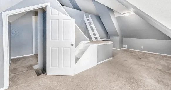 A room whose door is cut on a slant at the top, in front of a set of stairs that lead to a small landing and another set of stairs that appear to lead to a crawl space. There is an additional ladder that also leads to the crawl space. The room is full of 45-degree angles on the walls and ceiling.