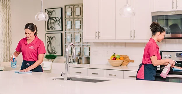 Two women in pink smiling and cleaning a kitchen.