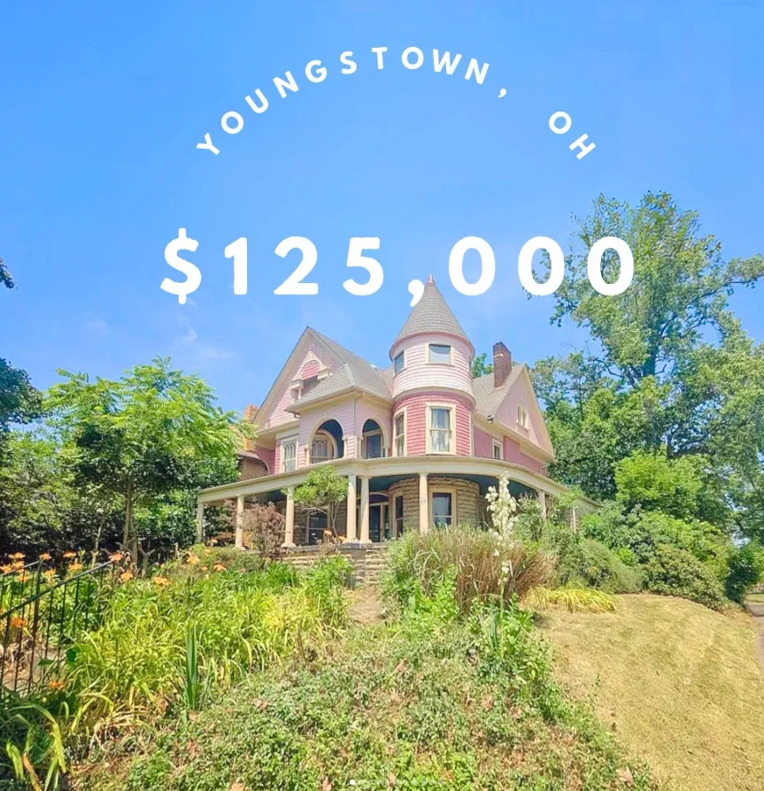 A pink Victorian mansion surrounded by grass and flowers.