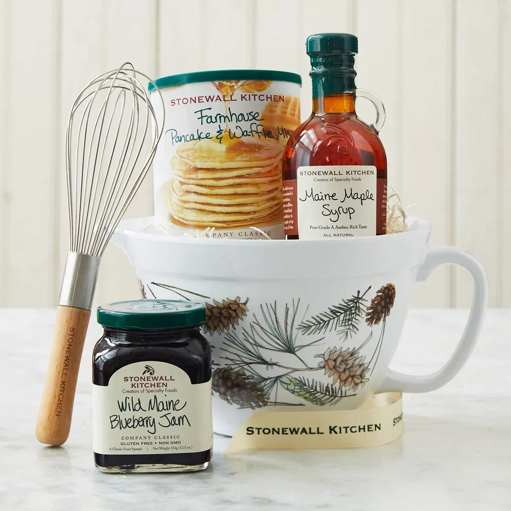 A mixing bowl with a pine design. Inside the bowl is a bottle of syrup and pancake and waffle mix. Next to the bowl is a jam jar and whisk.