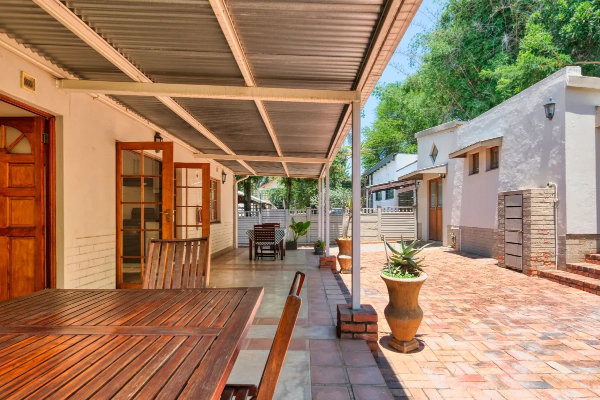 A stone patio off a home with wooden tables, chairs, and potted plants.